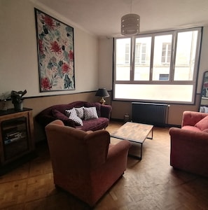 Un salon comprenant un canapé, un fauteuil et une table basse. Une fenêtre offre une vue sur les bâtiments à l’extérieur.