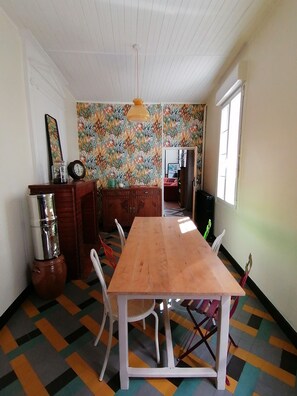 Un espace repas comprenant une table en bois, des chaises blanches, un mur à motifs et un plafond avec des poutres en bois.