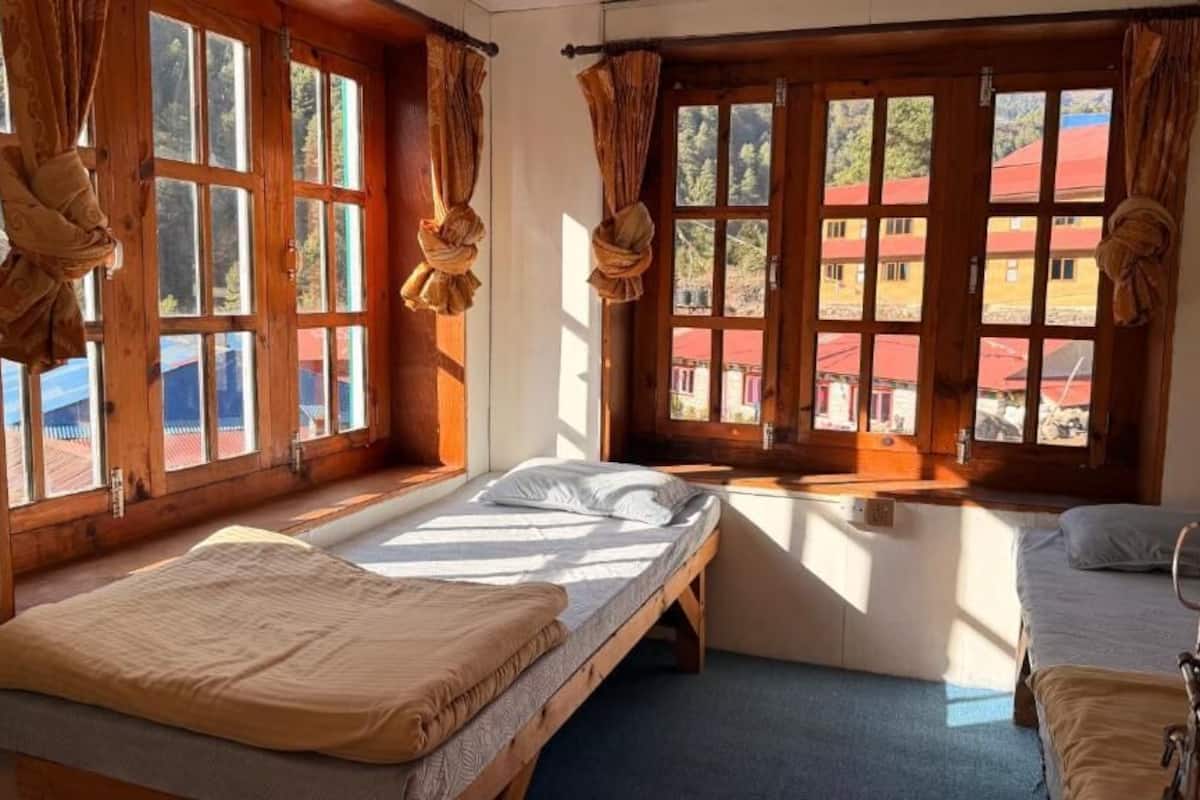A room with two beds, large windows with curtains, and a view of buildings outside.
