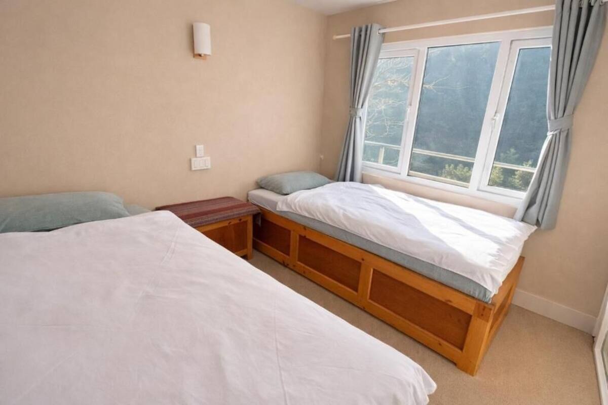 A bedroom with two beds, a window with curtains, a wall-mounted light, and a small bedside table.