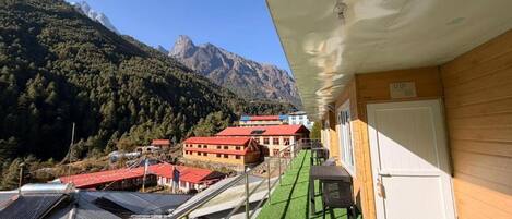 Habitación de hotel con balcón que da a un paisaje montañoso, un edificio de techo rojo al fondo y un césped artificial verde.