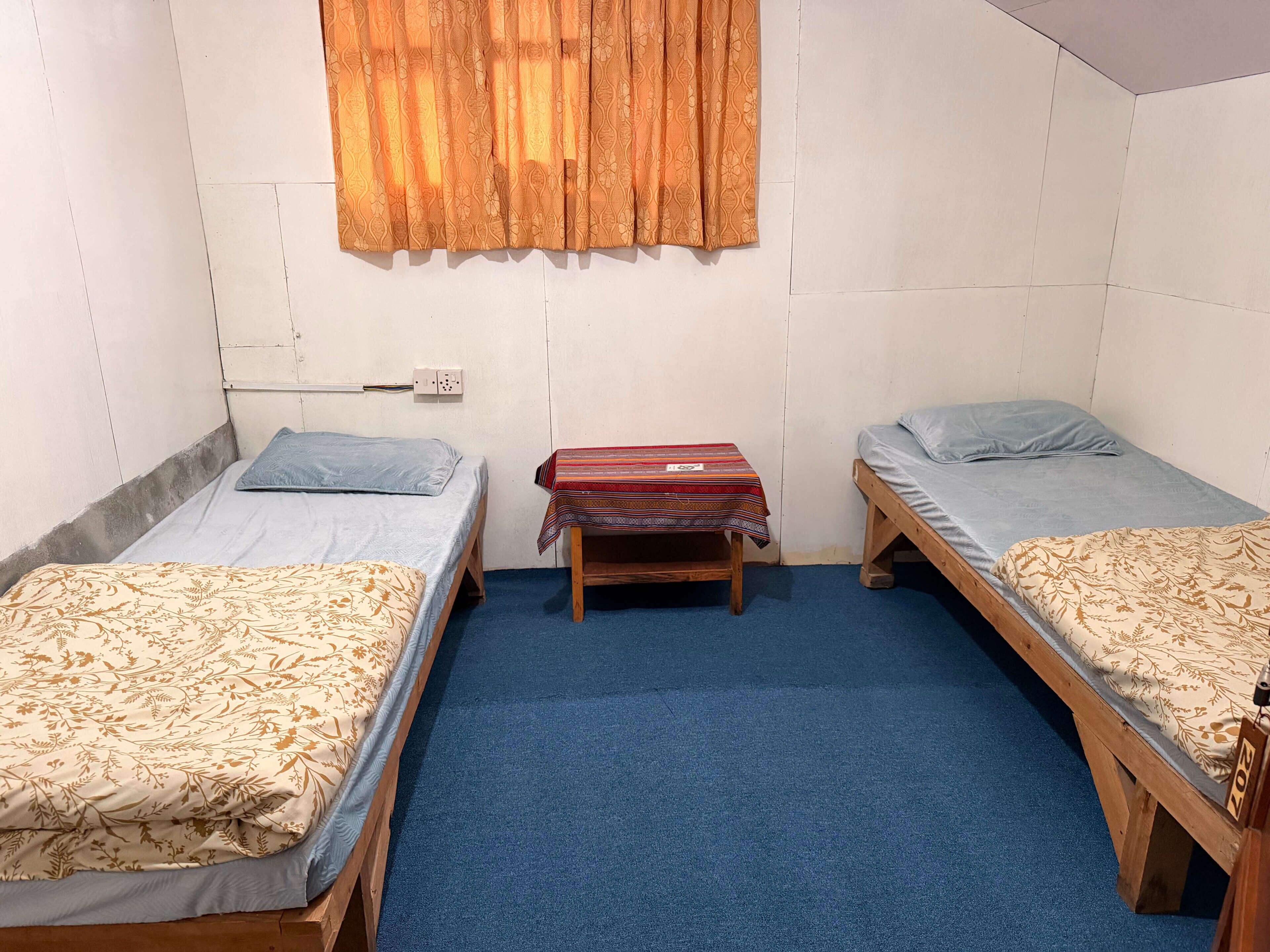 A dormitory room with two single beds, a small wooden table, a power outlet, and a window with curtains.