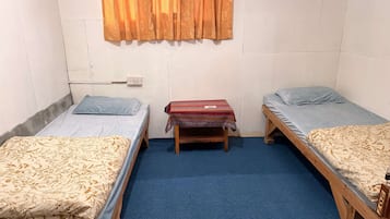 A dormitory room with two single beds, a small wooden table, a power outlet, and a window with curtains.