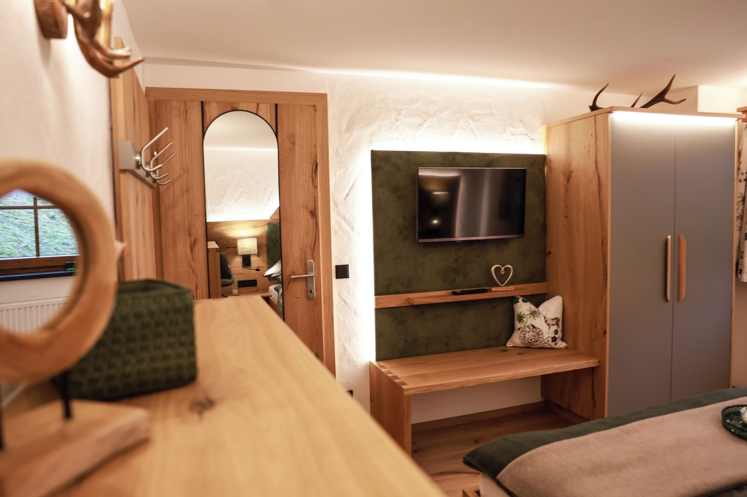 Ein Zimmer mit einem Holztisch, einem an der Wand montierten Fernseher, einem Bett mit grüner Decke und einem Kleiderschrank mit Spiegel.