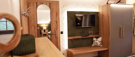 Ein Zimmer mit einem Holztisch, einem an der Wand montierten Fernseher, einem Bett mit grüner Decke und einem Kleiderschrank mit Spiegel.