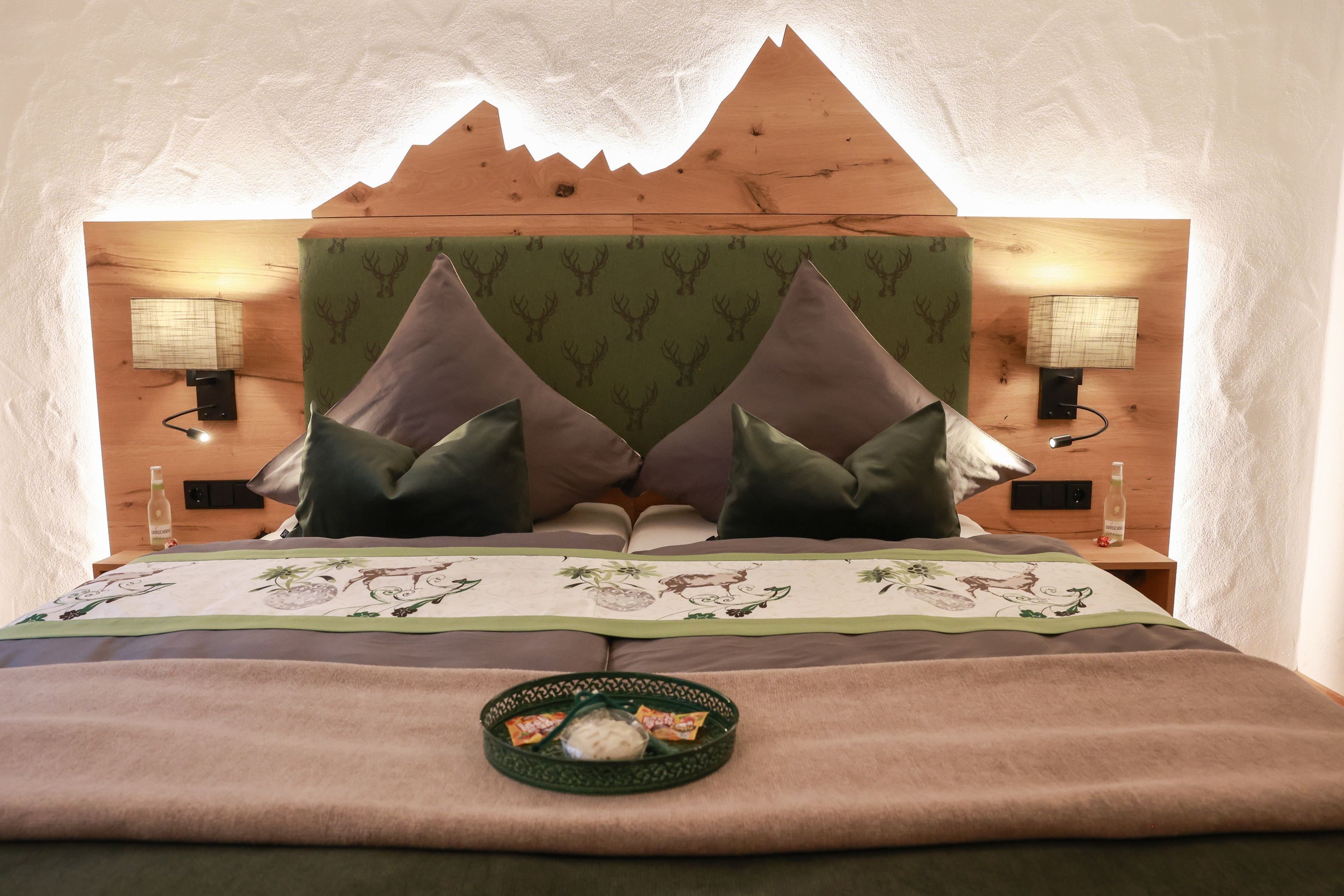 A bedroom with a mountain-shaped headboard, two bedside lamps, a bed with a floral bedspread, and a tray with snacks.