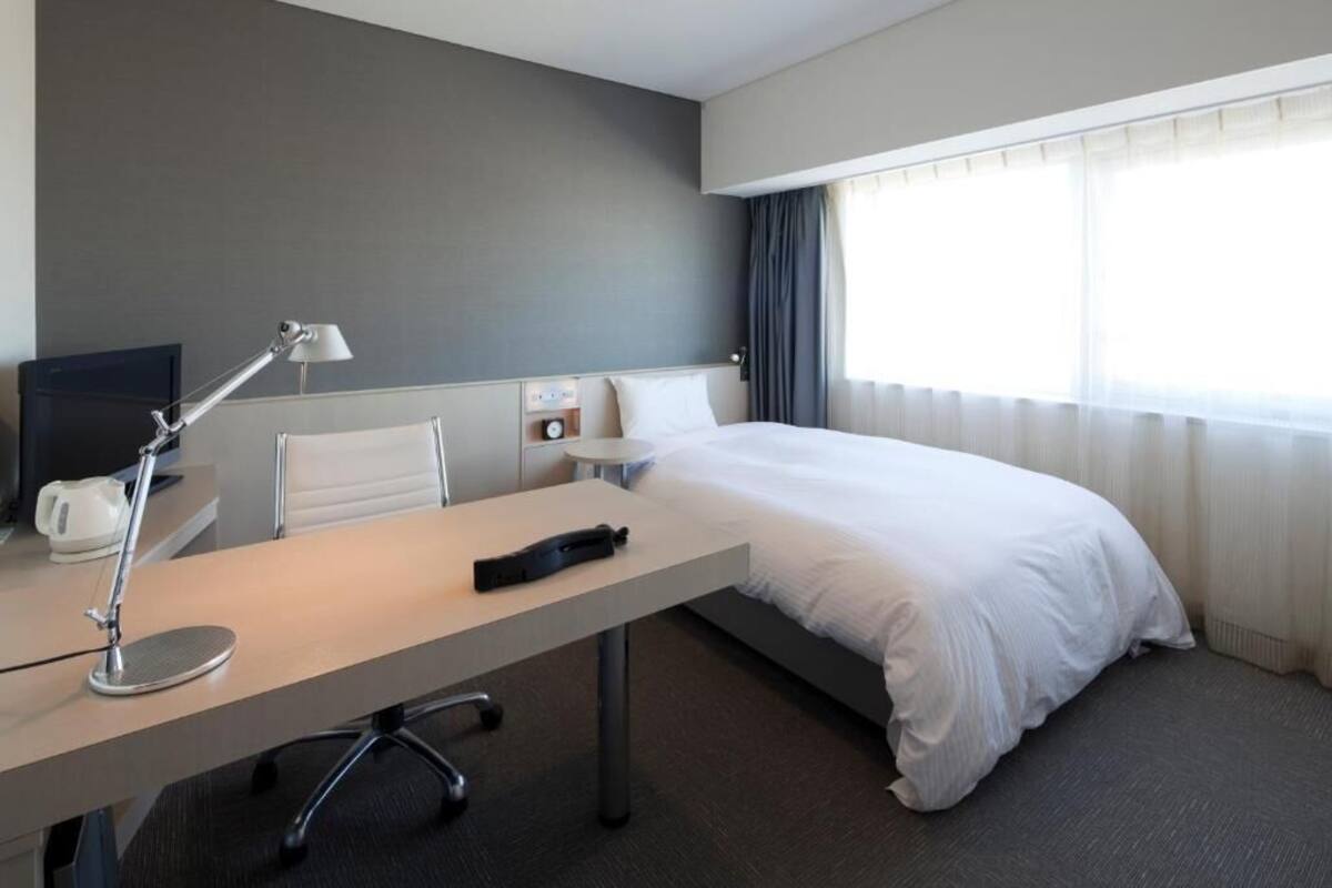 Habitación de hotel con cama, escritorio con lámpara, televisión y ventana con cortinas.
