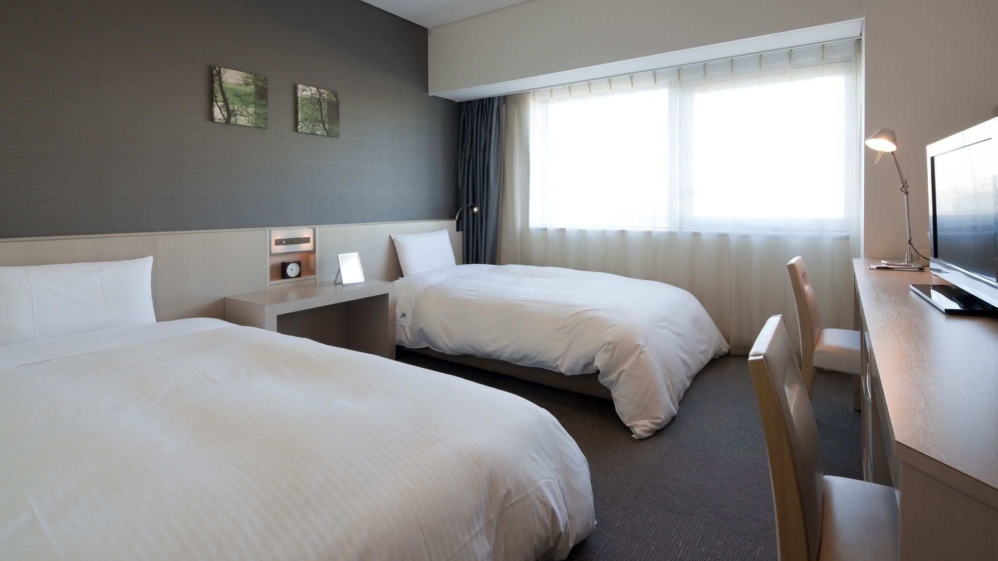 A hotel room with two beds, a desk with a lamp, a TV, and artwork on the wall.