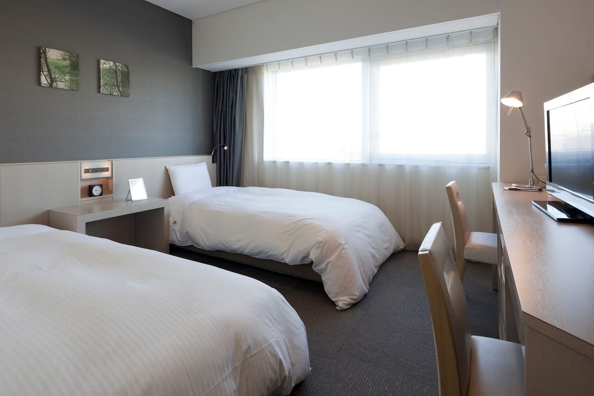 Habitación de hotel con dos camas, un escritorio con lámpara, televisor y obras de arte en la pared.