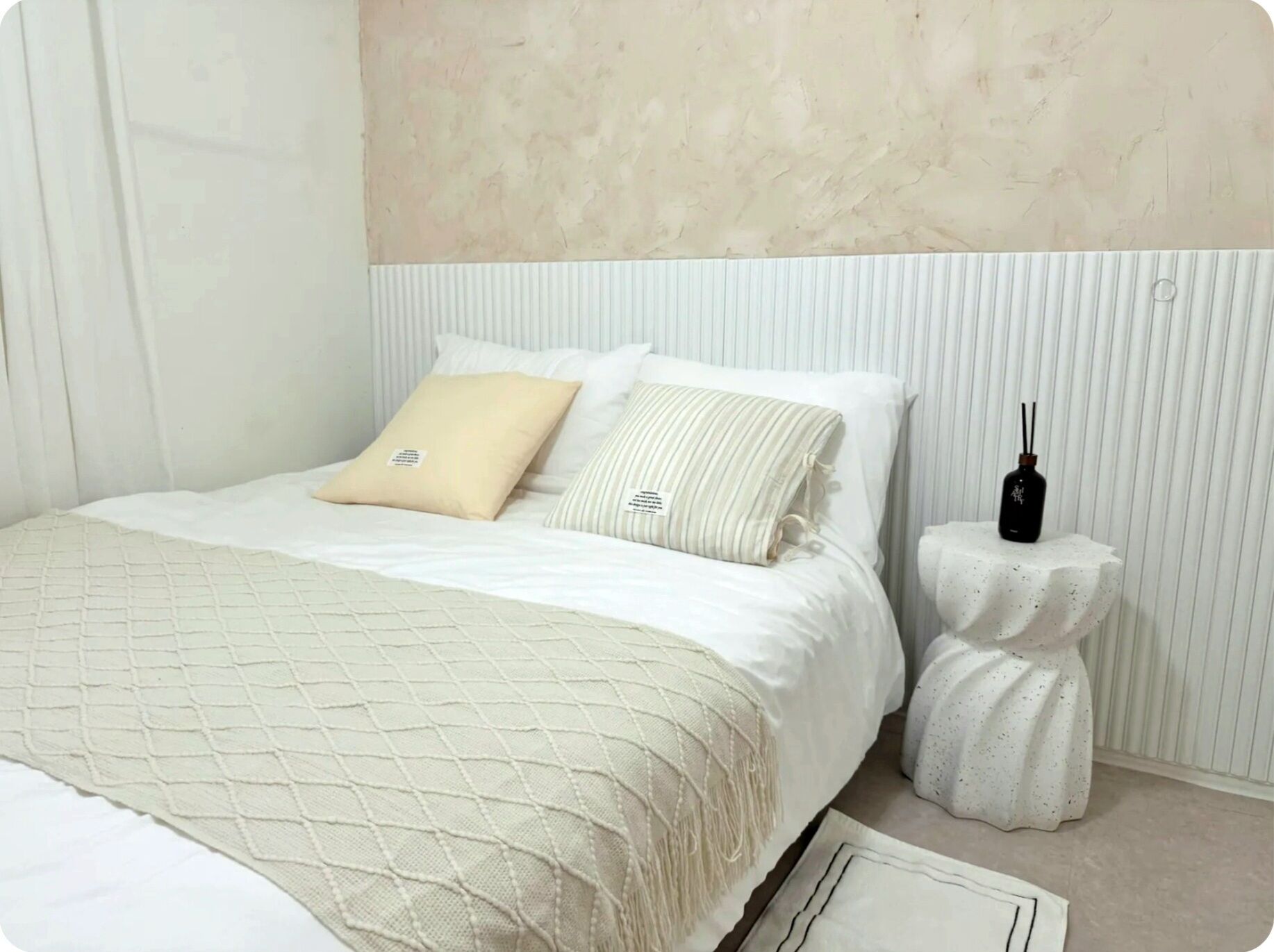 A bed with a quilted cover, two pillows, a bedside table with a black object, and a white wall with vertical lines.