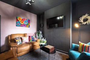 A living room with a brown leather sofa, a blue armchair, a colorful floral painting, a small wooden table, and a floor lamp. - Amen Hull Comfort 4BR Home | 2 Bathrooms | Central (Kingston upon Hull)