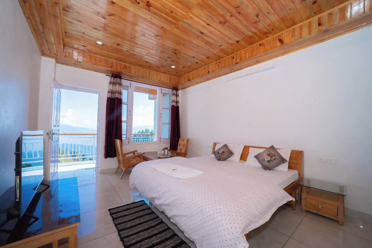 A bedroom with a wooden ceiling, a bed with pillows, a chair, a table, and a window with curtains.