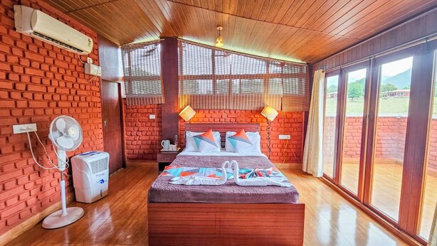 Une pièce avec un mur en briques, un lit recouvert d’une couvre-lit à motifs, un ventilateur sur pied, un radiateur et de grandes fenêtres donnant sur un beau paysage.