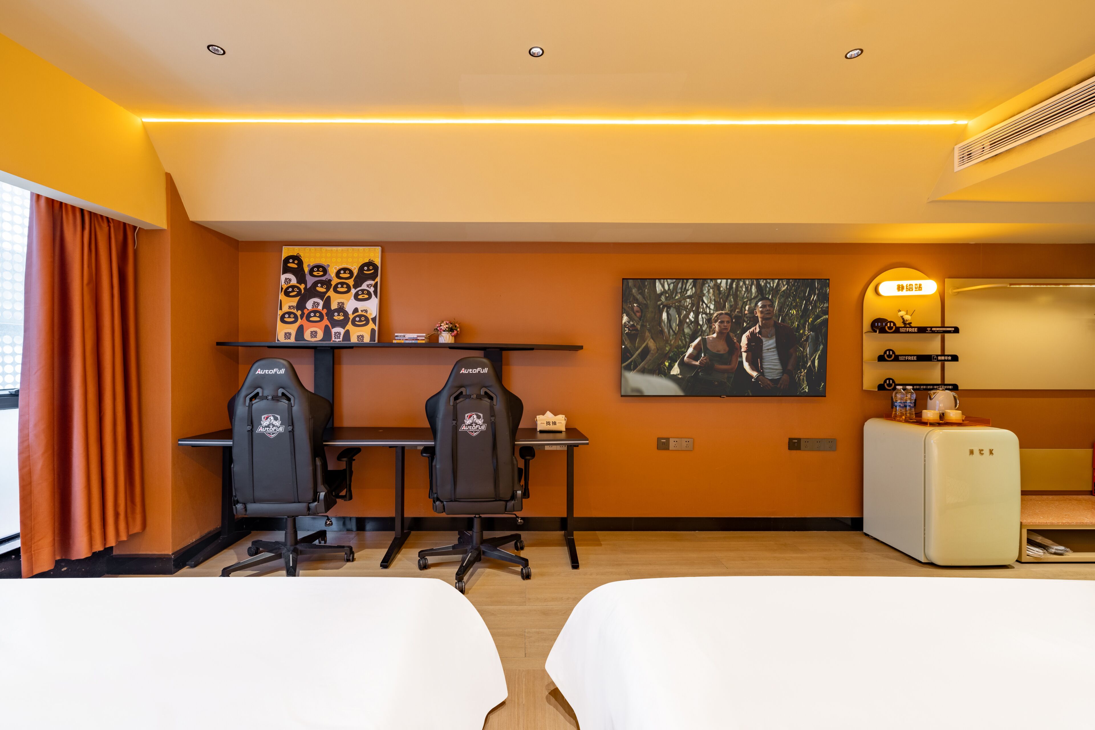 Camera d'albergo con due letti, una scrivania con due sedie da gaming, una televisione, un tavolino con un oggetto decorativo e una mensola a muro con una cornice.