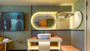Un baño moderno con tocador, un espejo grande y un televisor mostrando una escena de película.