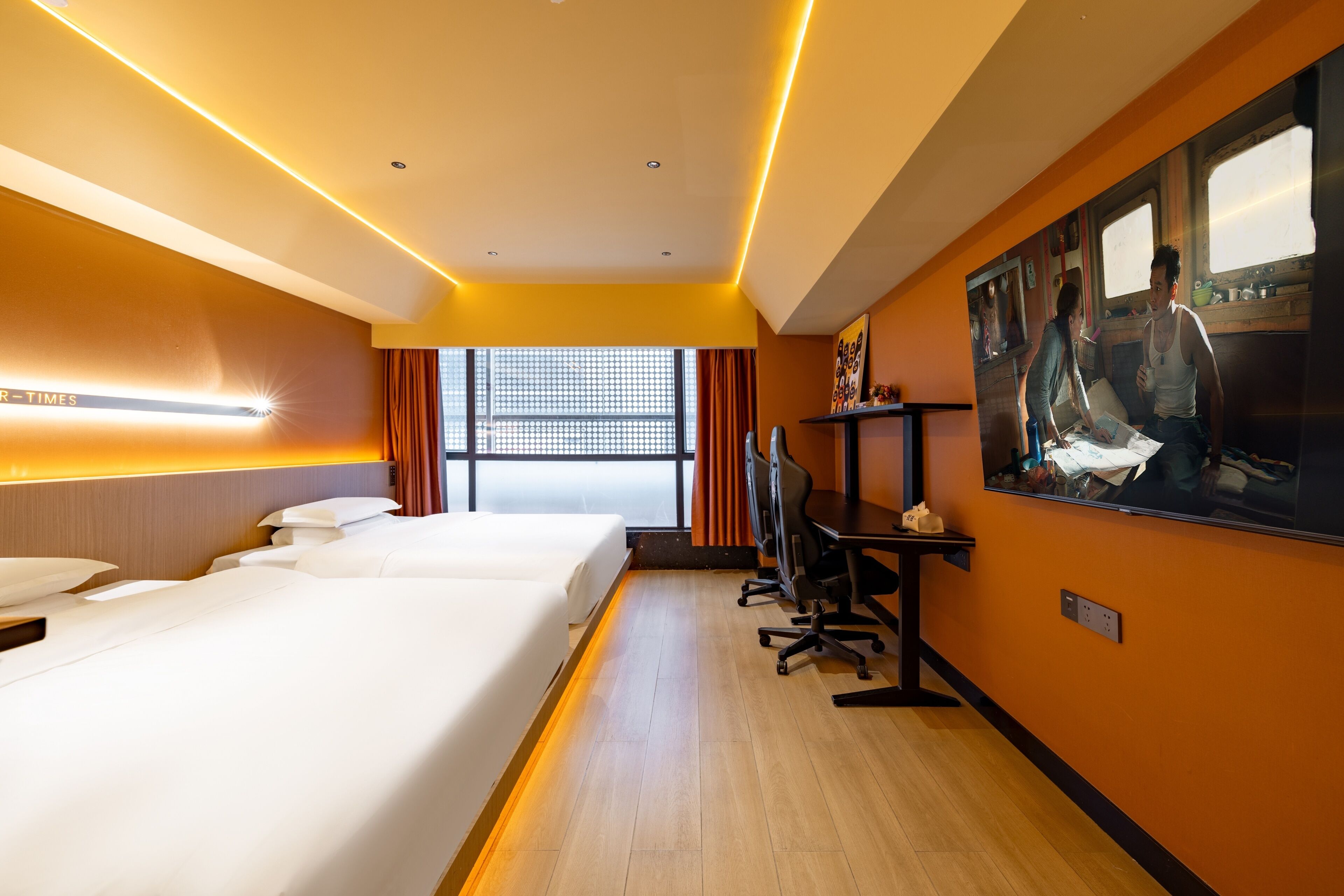 Ein Hotelzimmer mit zwei Betten, einem Schreibtisch, einem Stuhl, einem Fernseher und einem großen Fenster mit Vorhängen.
