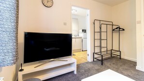 A modern living room with a flat-screen TV on a white stand, a coat rack, and a kitchen visible through an open door. - One Bed Apartment in Wellington (Telford and Wrekin)