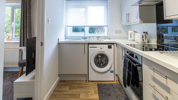 A compact kitchen with a washing machine, a built-in oven, and a window with a view of greenery. - One Bed Apartment in Wellington (Telford and Wrekin)