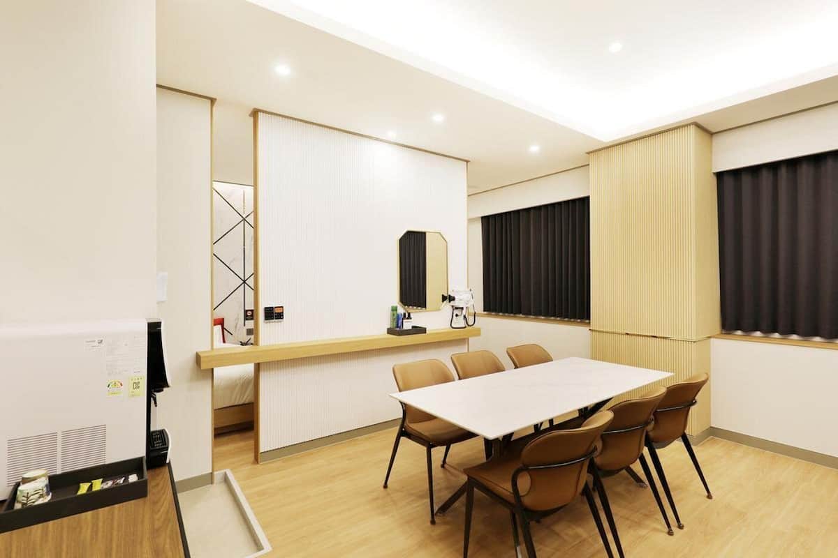 A hotel room with a large bed, a desk with a computer, a chair, a table with chairs, and a window with curtains.