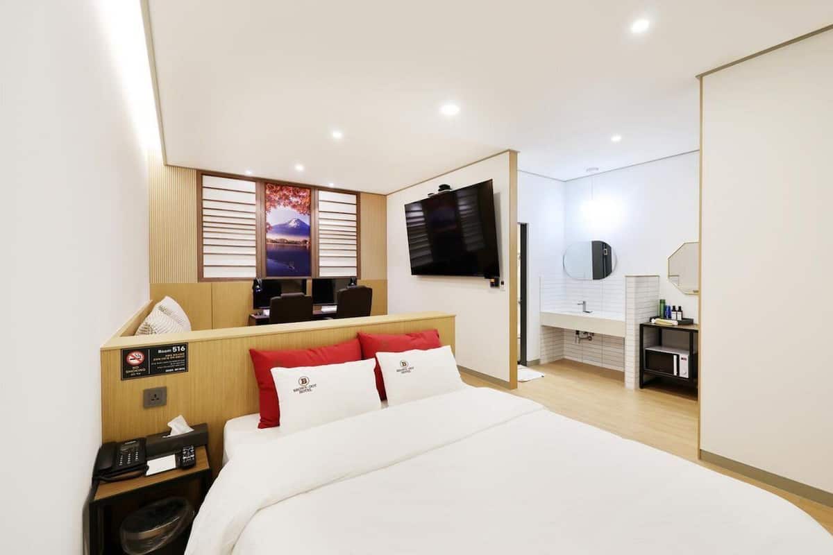 A hotel room with a bed, a desk with a computer, a TV, a bathroom with a mirror, and a view of a mountain.