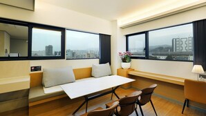 A modern hotel room with a sofa, dining table, chairs, a small side table with a flower vase, and large windows offering city views. - Hotel Brown Dot (Dongducheon Central Station) (Dongducheon)