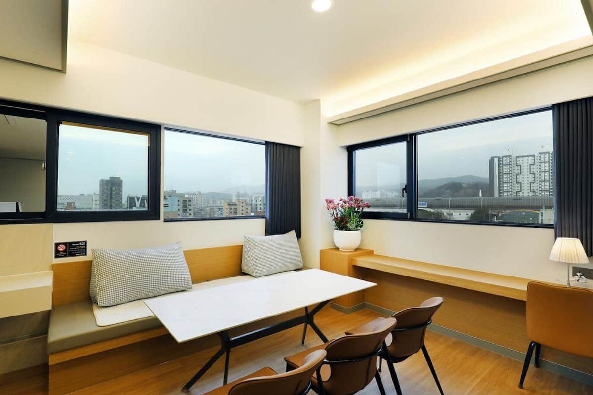 Une chambre d’hôtel moderne équipée d’un canapé, d’une table à manger, de chaises, d’une petite table d’appoint avec un vase de fleurs et de grandes fenêtres offrant une vue sur la ville.
