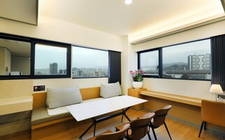 A modern hotel room with a sofa, dining table, chairs, a small side table with a flower vase, and large windows offering city views.