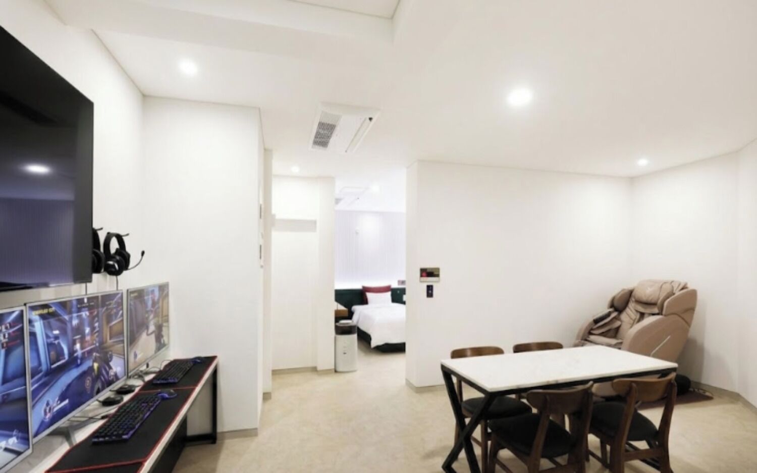 A modern hotel room with a flat-screen TV, gaming monitors, a massage chair, and a dining table.