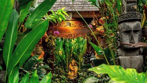 Un sentier de jardin mène à un bâtiment dont le toit est recouvert de chaume, entouré d’une végétation tropicale luxuriante et d’une statue en bois.