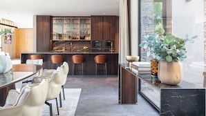 A modern kitchen with dark wood cabinets, a marble countertop, and a dining area with leather chairs. - New build villa right on the beach with tropical garden and Bali feeling (Cala Vinyes)