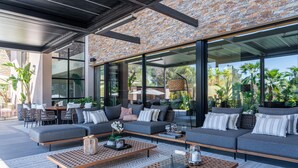 A modern outdoor seating area with sofas, tables, and chairs, featuring a stone wall and large windows. - New build villa right on the beach with tropical garden and Bali feeling (Cala Vinyes)