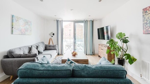 A modern living room with a grey sofa, a teal sofa, a wooden coffee table, and a potted plant. - Beautiful 1 Bedroom Waterfront Apartment with parking in Gloucester Docks (Gloucester)