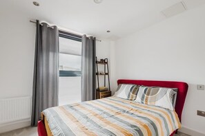 Une chambre à coucher avec un lit, des rideaux, une table de chevet et une vue sur l’extérieur.