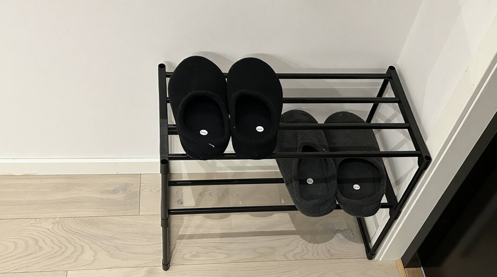 Un porte-chaussures en métal noir, avec quatre paires de pantoufles noires, est placé contre un mur blanc et sur un plancher en bois.