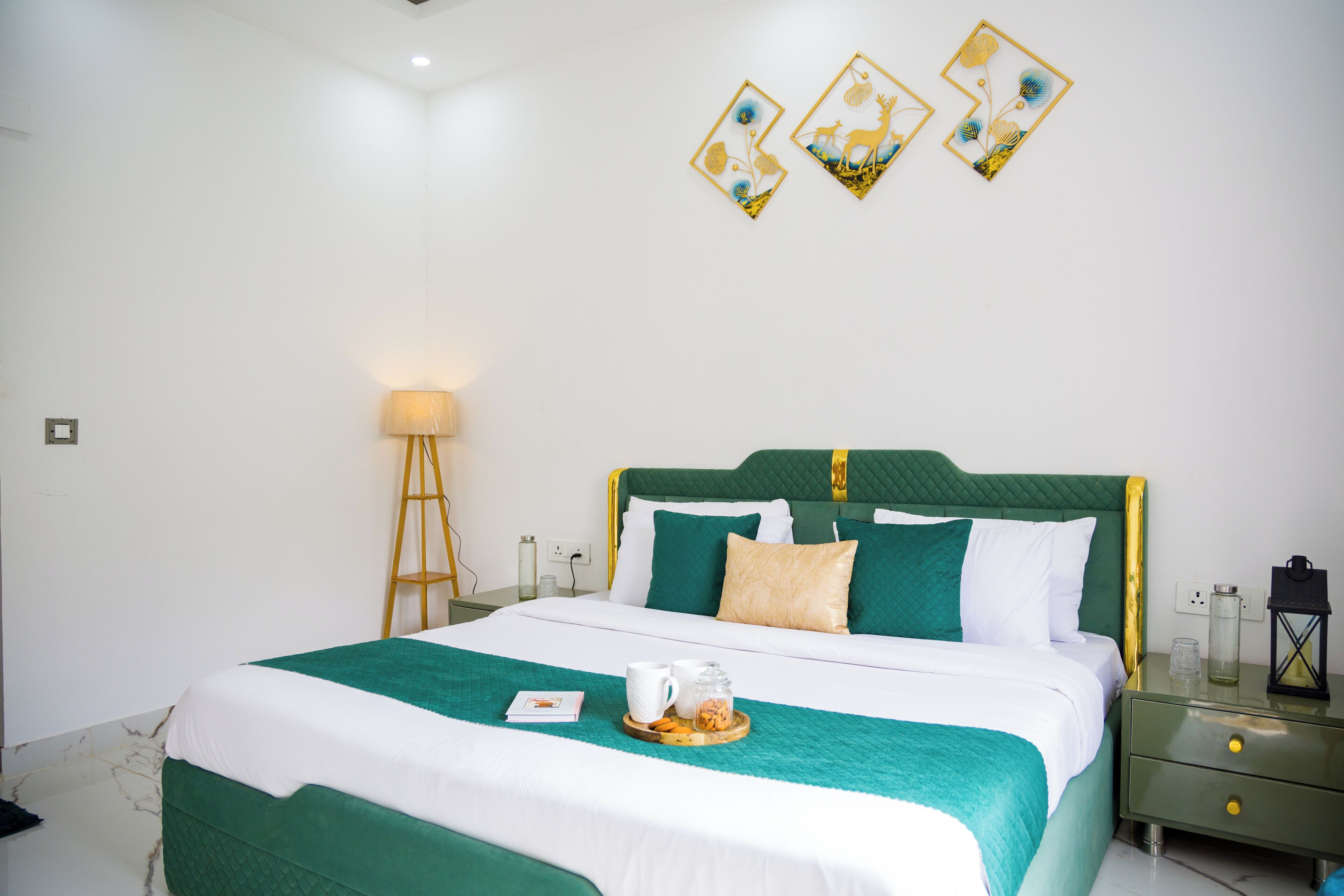 Un lit bien fait, recouvert d’une couvre-lit verte et blanche, une table de chevet avec un verre et une bougie, et des œuvres d’art accrochées au mur au-dessus du lit.
