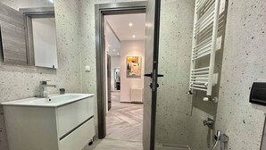 Un baño moderno con un lavamanos blanco, espejo y retrete, una puerta de cristal para la ducha, y un pasillo que conduce a una sala de estar.