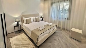 Un dormitorio moderno con una cama grande, lámparas de noche, una ventana con cortinas translúcidas y un pequeño otoman.