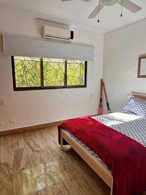 Un dormitorio con cama, ventana con vista a árboles, unidad de aire acondicionado y ventilador de techo.