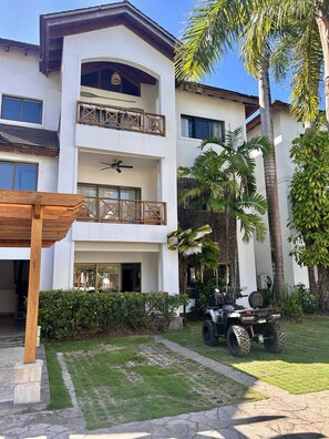 Un edificio de varios pisos con balcones, un patio cubierto y un césped con un vehículo.