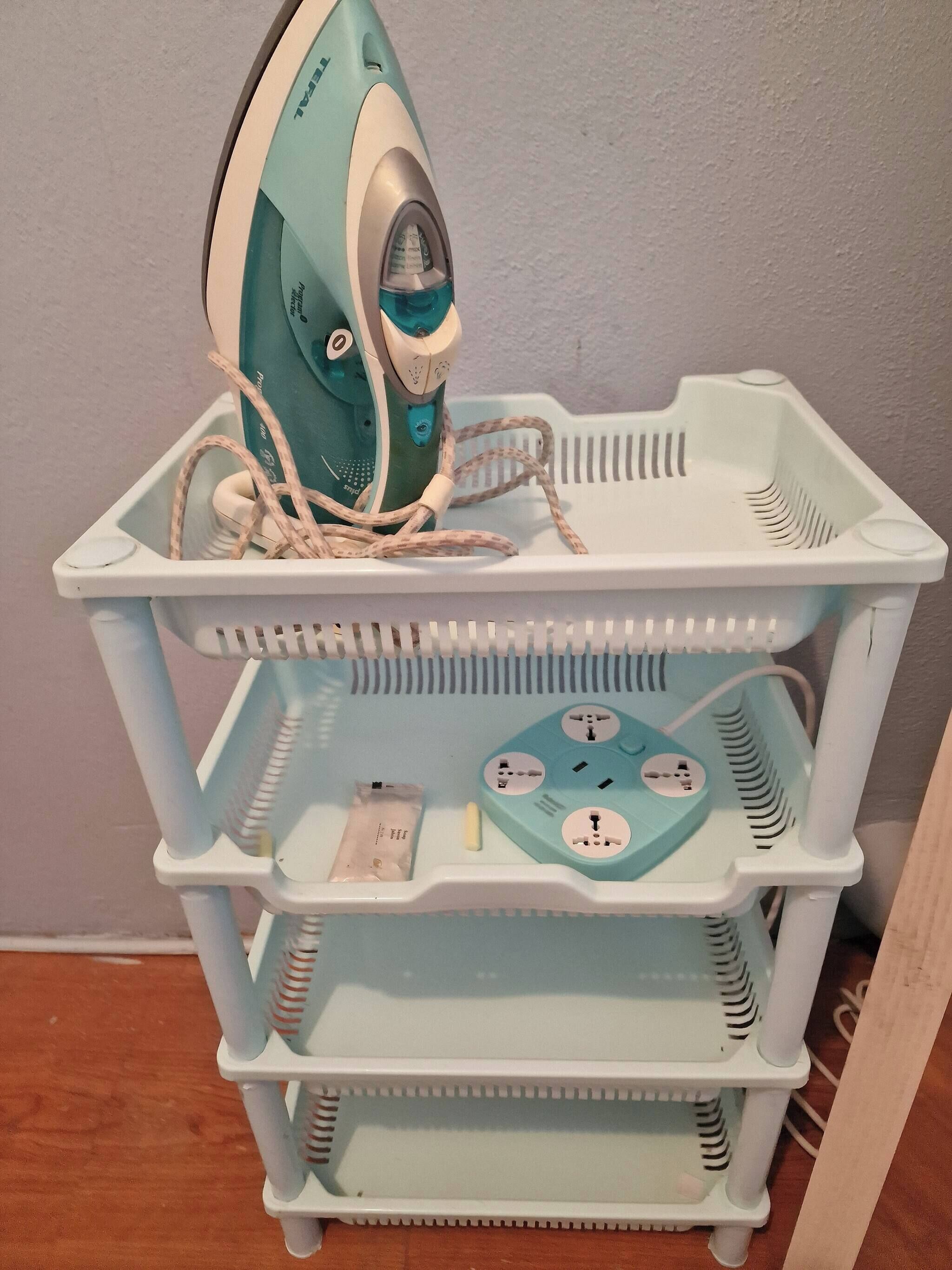 A white plastic storage cart with multiple compartments, including a top shelf holding an iron and tangled cords.