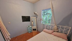 A bedroom with a bed, a window with a view, a wall-mounted fan, and a small shelf.