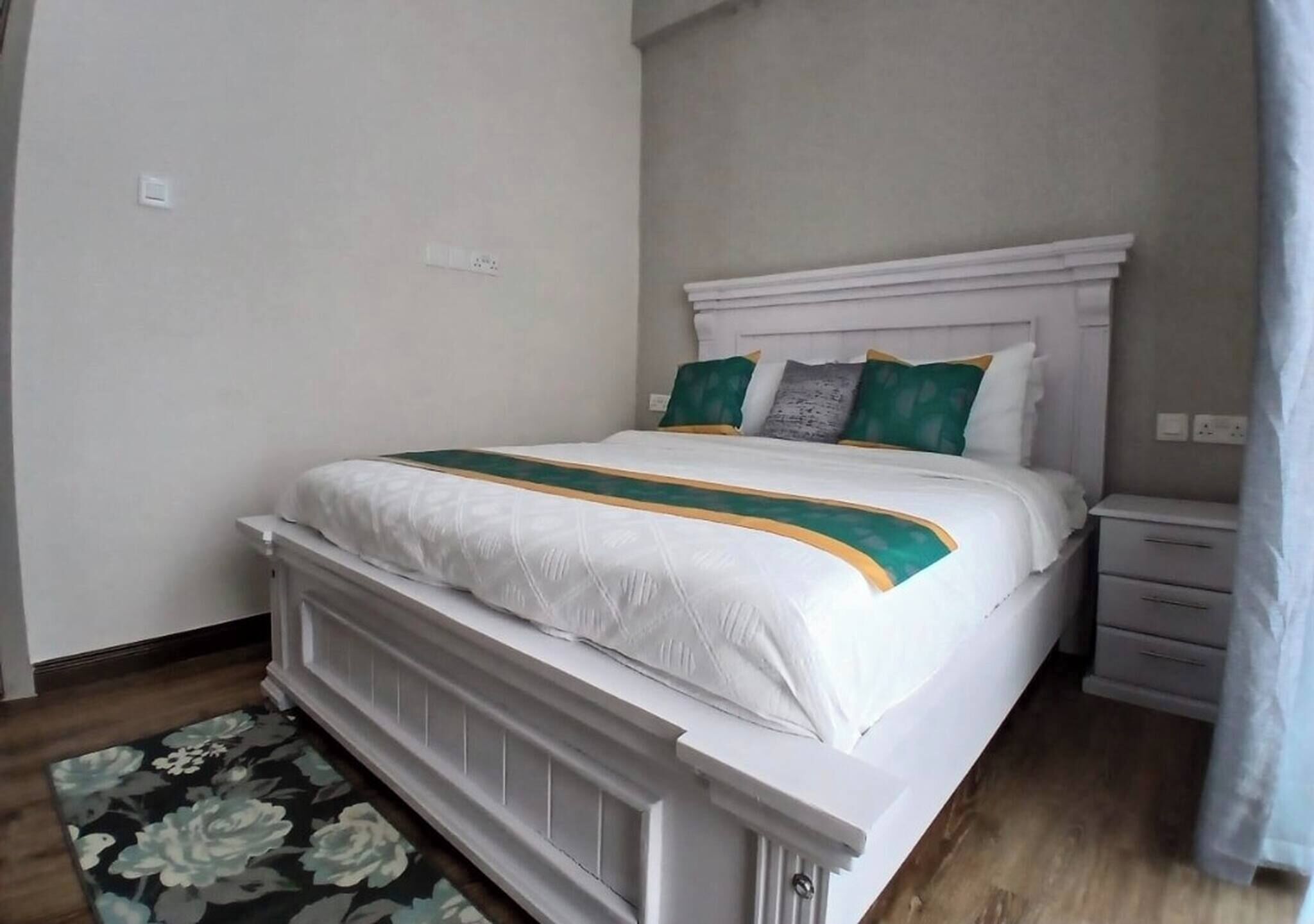 Una cama bien tendida con un cabecero blanco, una mesita de noche con cajones y una alfombra estampada en el suelo.