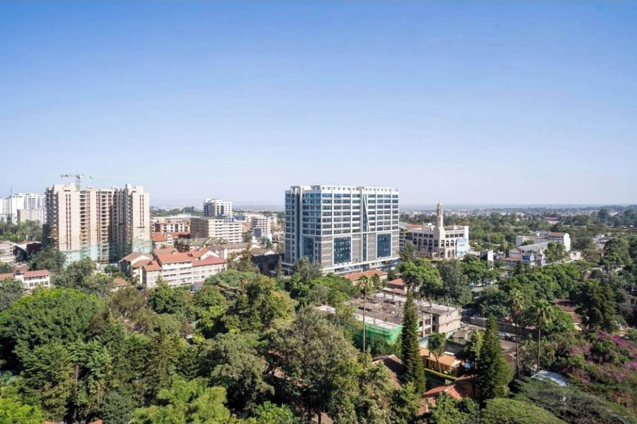 A cityscape with modern buildings, dense greenery, and a clear sky.