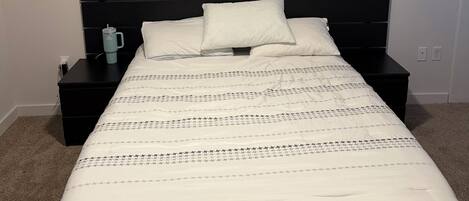 A neatly made bed with a white comforter and pillows, a black headboard, a bedside table with a mug, and a power outlet on the wall.