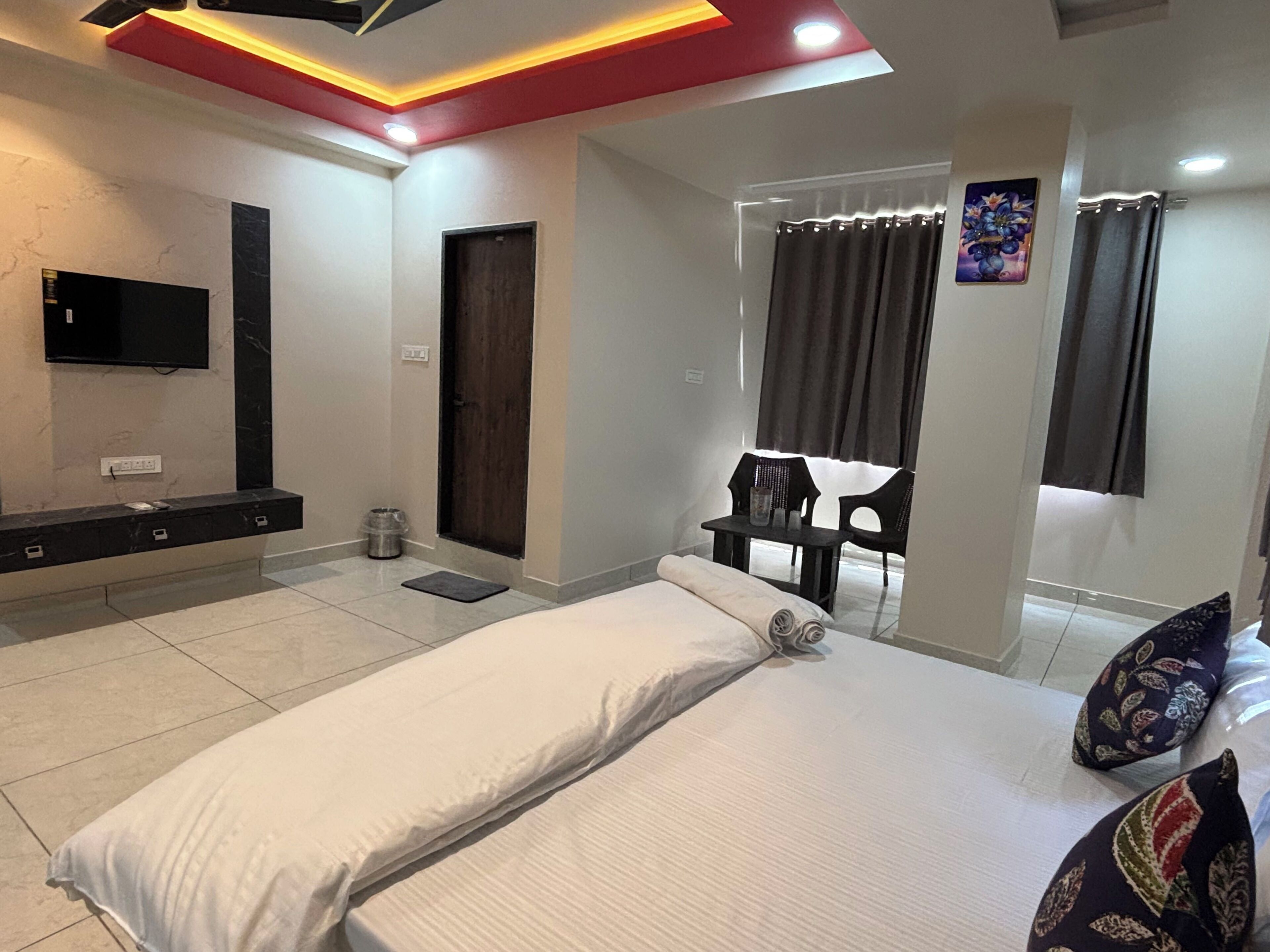 A hotel room with a bed, a TV mounted on the wall, a small table with chairs, and a window with curtains.