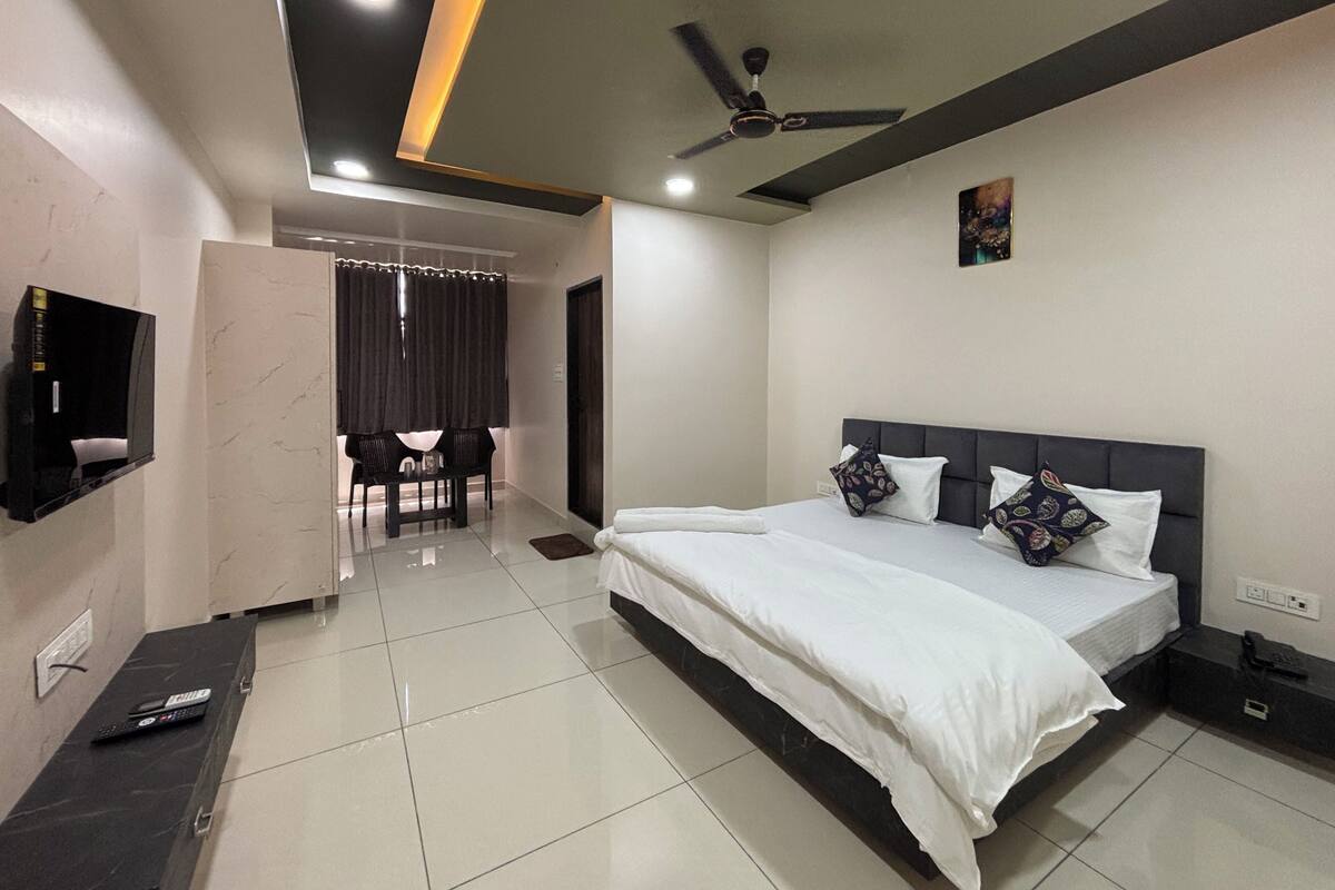 A modern hotel room with a large bed, a ceiling fan, a flat-screen TV mounted on the wall, and a small seating area with a table and chairs.