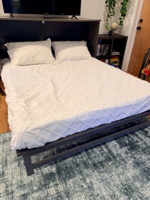 A bed with a quilted white cover, a dark headboard, and a grey carpet.