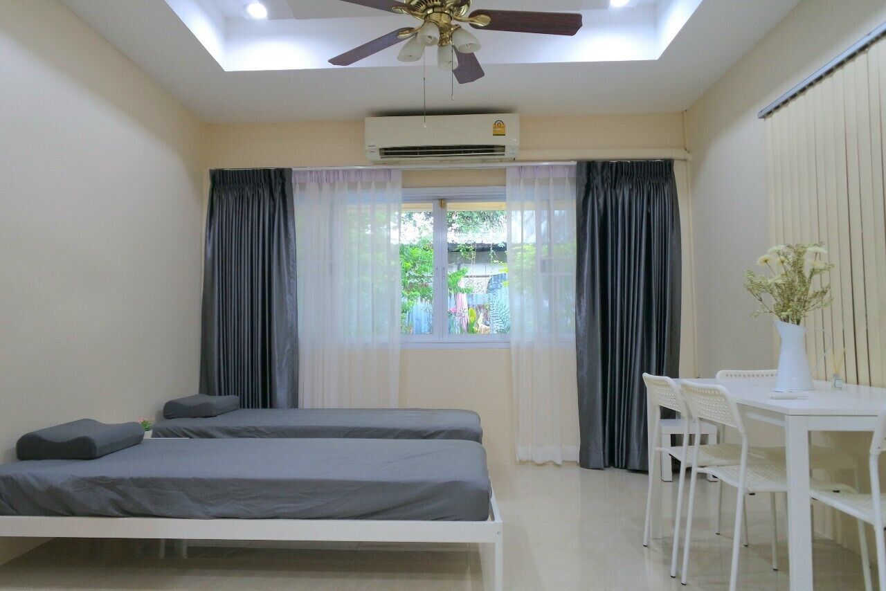 A bedroom with two beds, a ceiling fan, an air conditioning unit, a window with curtains, and a small table with chairs.