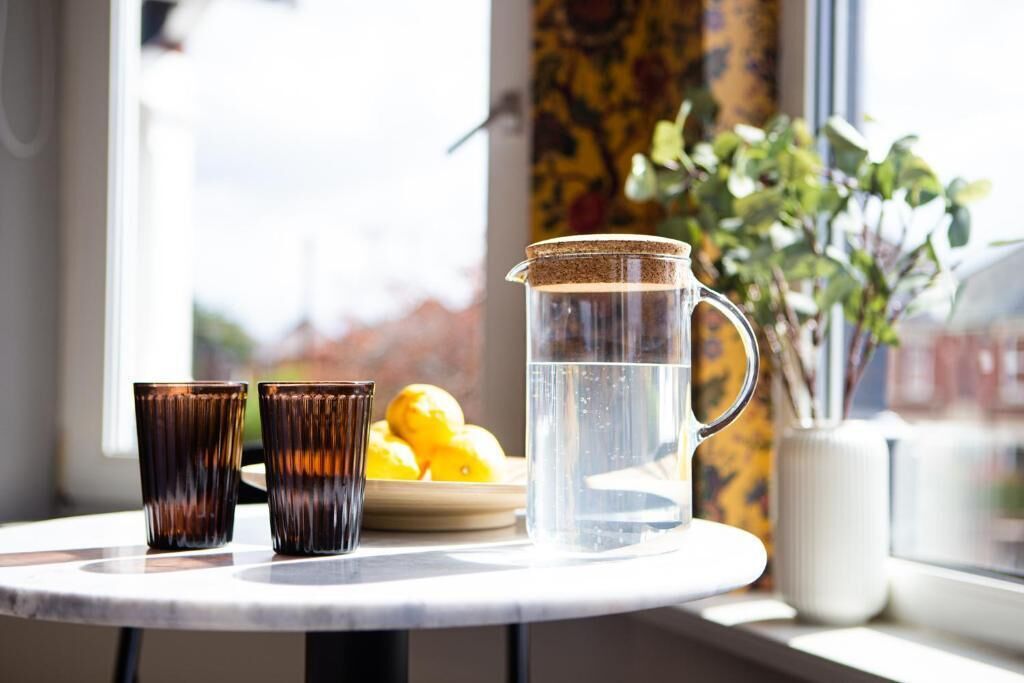 Una mesa con una jarras de agua, dos vasos y un plato de limones junto a una ventana.
