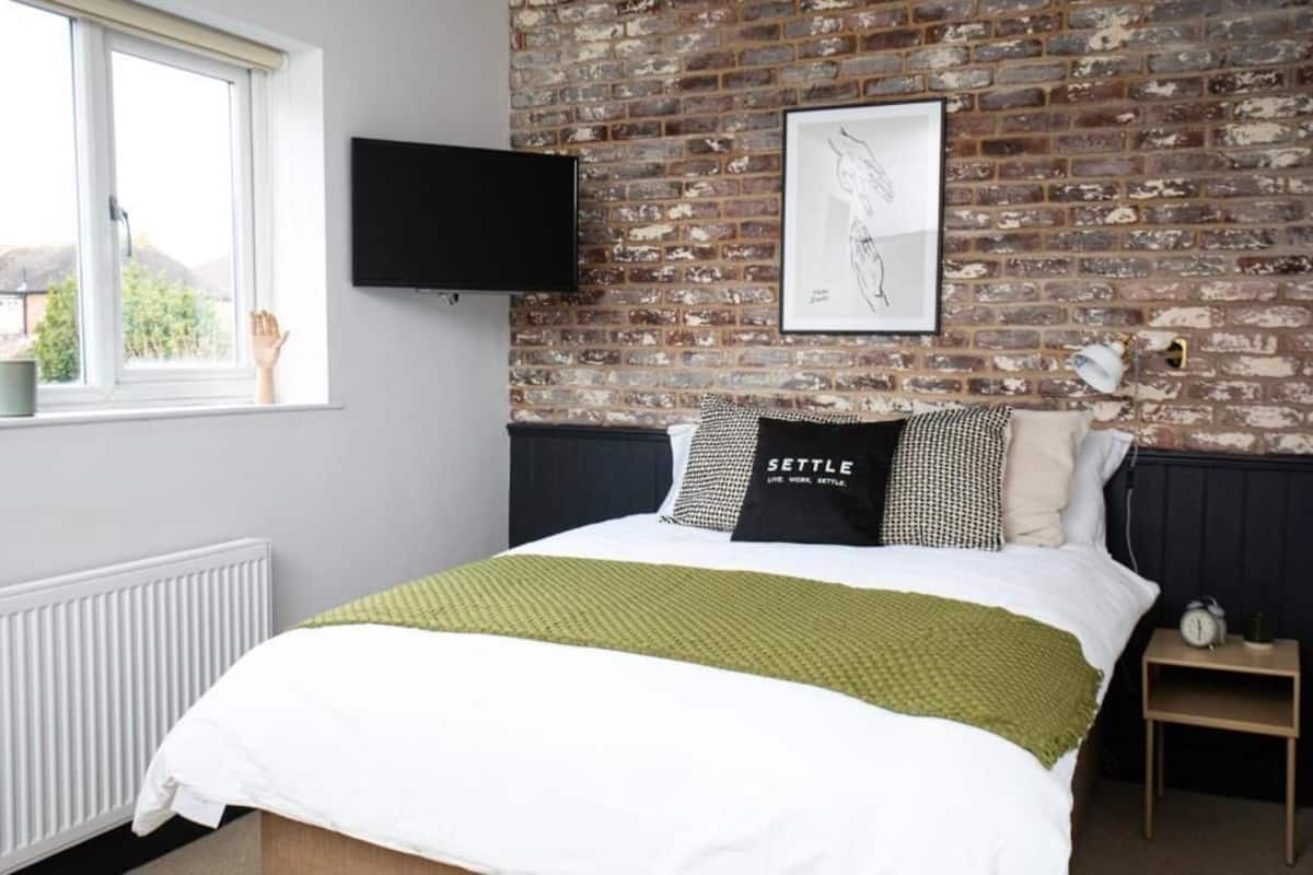 A bedroom with a brick wall, a bed with a green blanket, a mounted TV, and a framed picture.